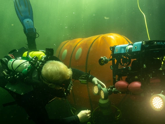 A scuba diver services an underwater research robot with an underwater camping tent in the background.