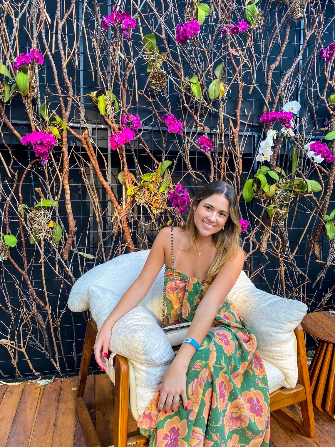 Maria smiling at the camera wearing a floral dress while sitting on a comfy chair. There are pretty vines with flowers in the background.