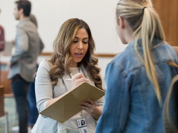 A healthcare worker writing notes on a clipboard while talking to another woman