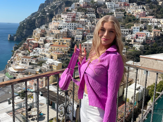 Ellie Moore standing at a viewing point in the Amalfi Coast in Italy. The view shows a blue ocean with a cliffside of colorful buildings in the greenery.