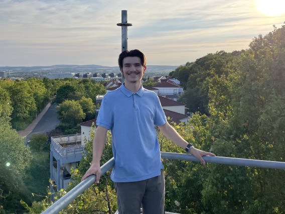 Jacob standing at a view point with a very green background. The sky in the background is blue with warm tones and looks like the sun is about to set. He is wearing a collared shirt with slacks.