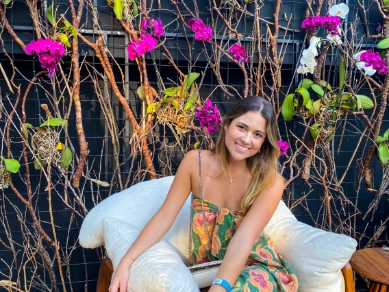 Maria smiling at the camera wearing a floral dress while sitting on a comfy chair. There are pretty vines with flowers in the background.