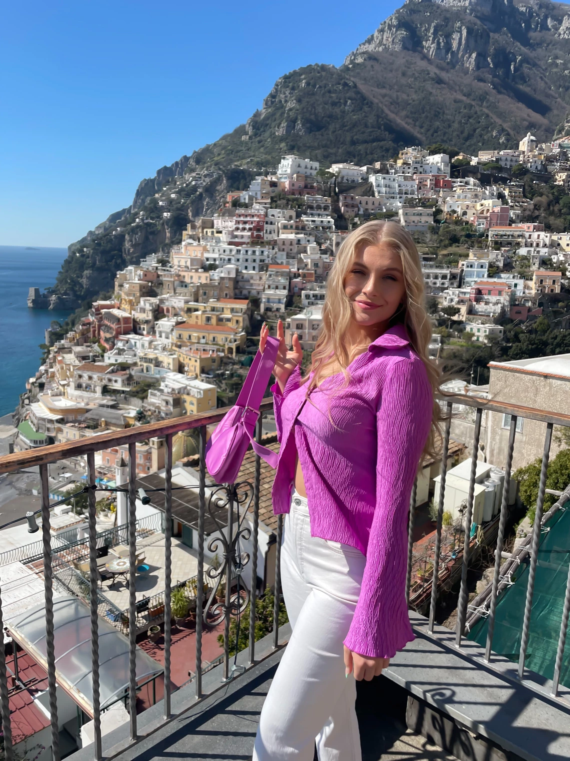 Ellie Moore standing at a viewing point in the Amalfi Coast in Italy. The view shows a blue ocean with a cliffside of colorful buildings in the greenery.