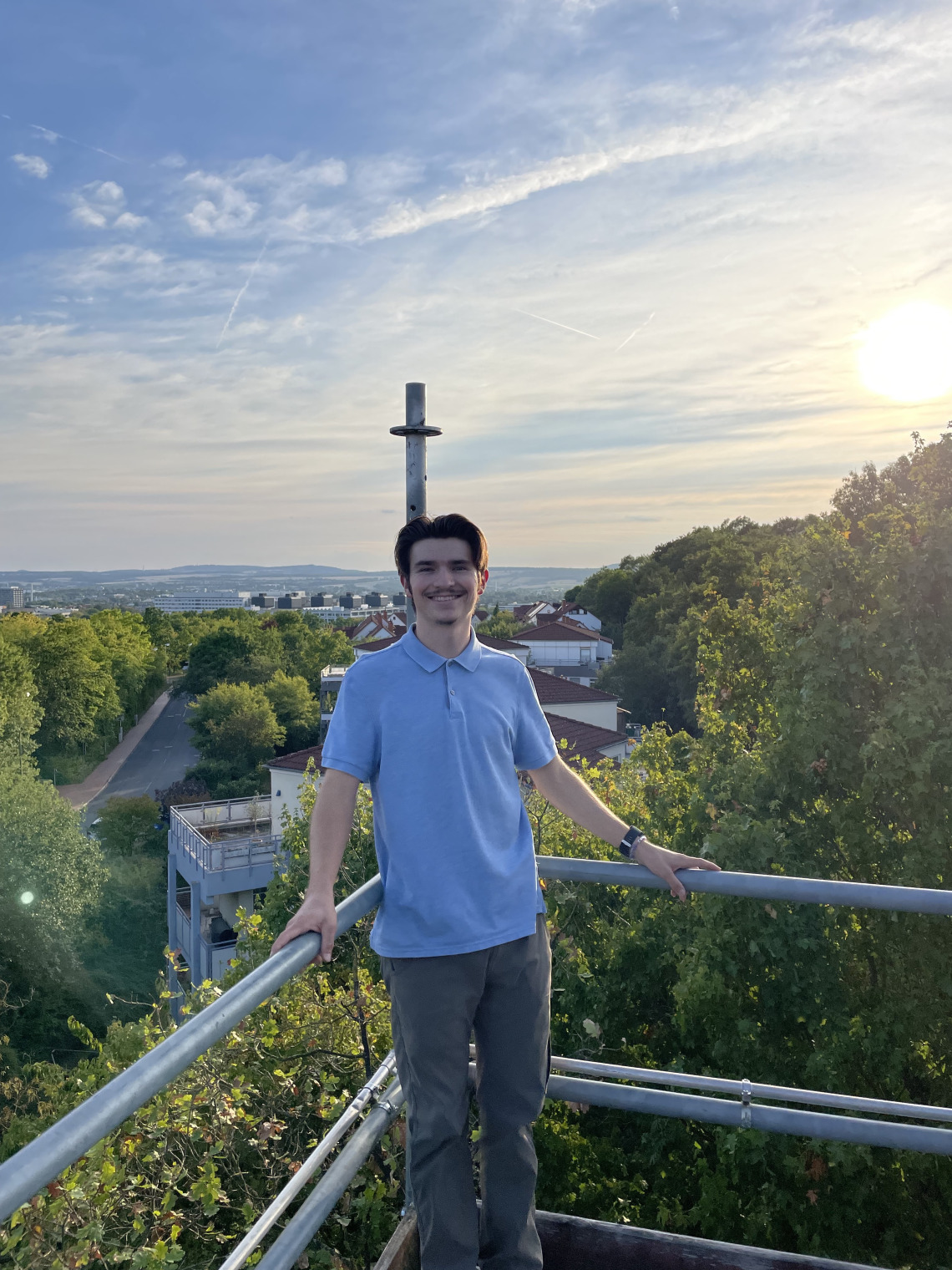 Jacob standing at a view point with a very green background. The sky in the background is blue with warm tones and looks like the sun is about to set. He is wearing a collared shirt with slacks.