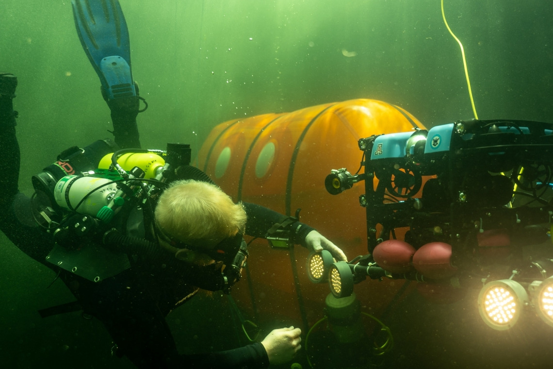 A scuba diver services an underwater research robot with an underwater camping tent in the background.